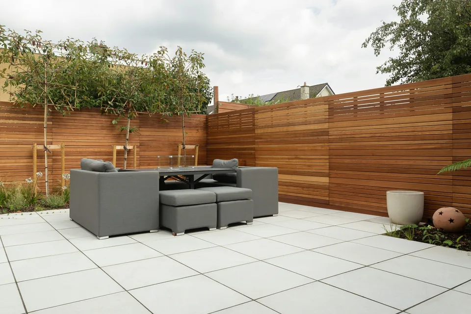 Ruhiger Gartensitzplatz mit hellen Terrassenplatten, gepflanzten Bäumen und horizontalem Holzsichtschutz.